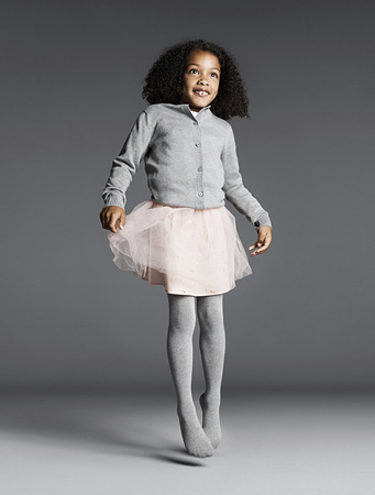 Ein fröhliches Mädchen springt in die Luft, gekleidet in eine graue FALKE Family Strumpfhose, einen rosa Tüllrock und eine graue Strickjacke.